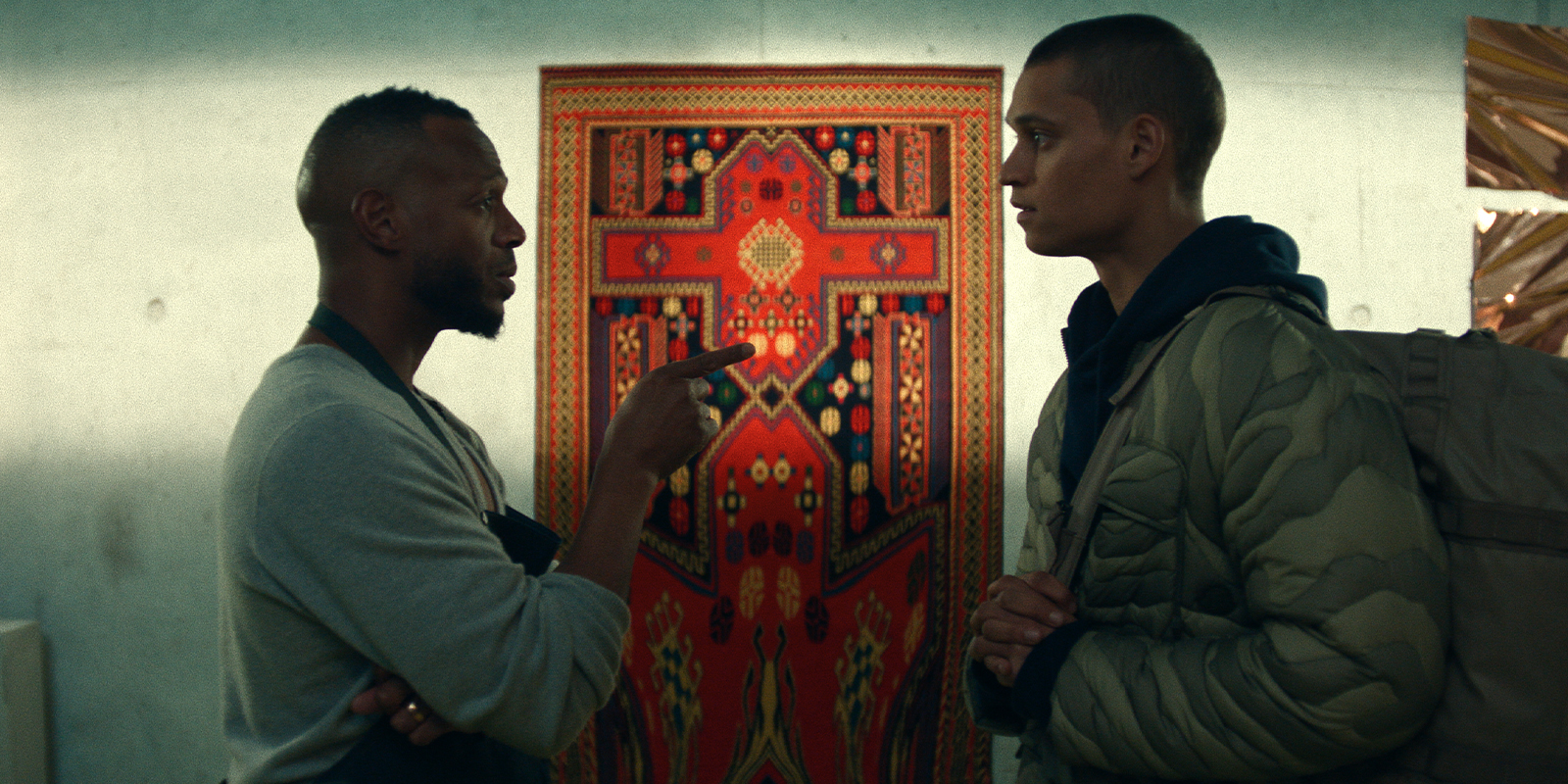 Two people stand facing each other indoors; one gestures while talking, the other listens with a serious expression. Behind them, a colourful, patterned rug hangs on the wall—cinematography reminiscent from Kira Kelly, BSC.