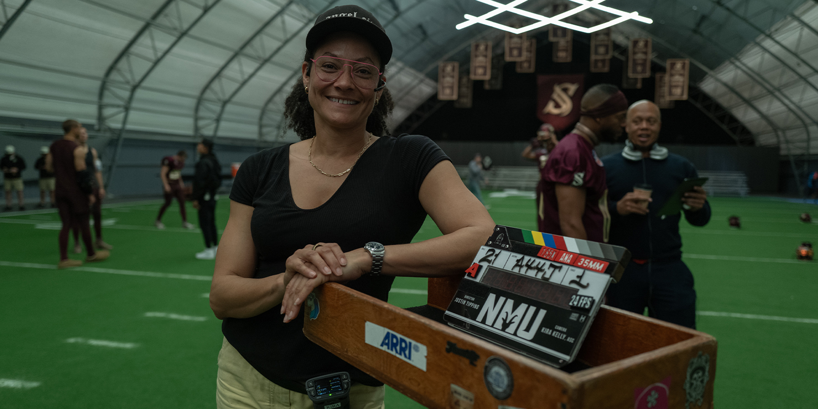 A smiling woman wearing glasses and a cap, possibly Kira Kelly, ASC, stands on an indoor sports pitch, leaning on a box holding a film clapperboard. People and sports banners can be seen in the background.