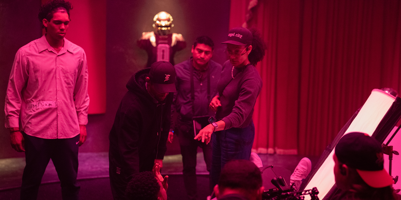 Under Kira Kelly’s dynamic lighting, a group collaborates on a dimly lit stage; red and purple hues surround them as Him, in a football kit, stands in the background whilst others engage in discussion up front.