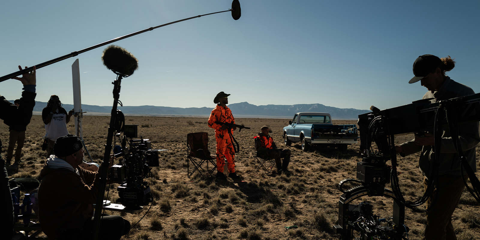 A film crew, led by Kira Kelly, ASC, shoots a scene outdoors on a dry, grassy plain with mountains in the distance. Two actors—one in bright orange kit, one sitting—face the camera as crew members operate equipment around him.