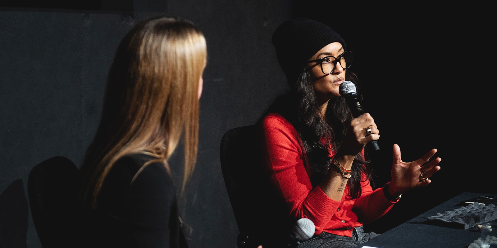 Two women sit at a table in conversation. One, wearing a red shirt, glasses, and a black beanie, gestures while speaking into a microphone during an event review. The other woman, with long blonde hair, faces her as they discuss Camerimage 2026.
