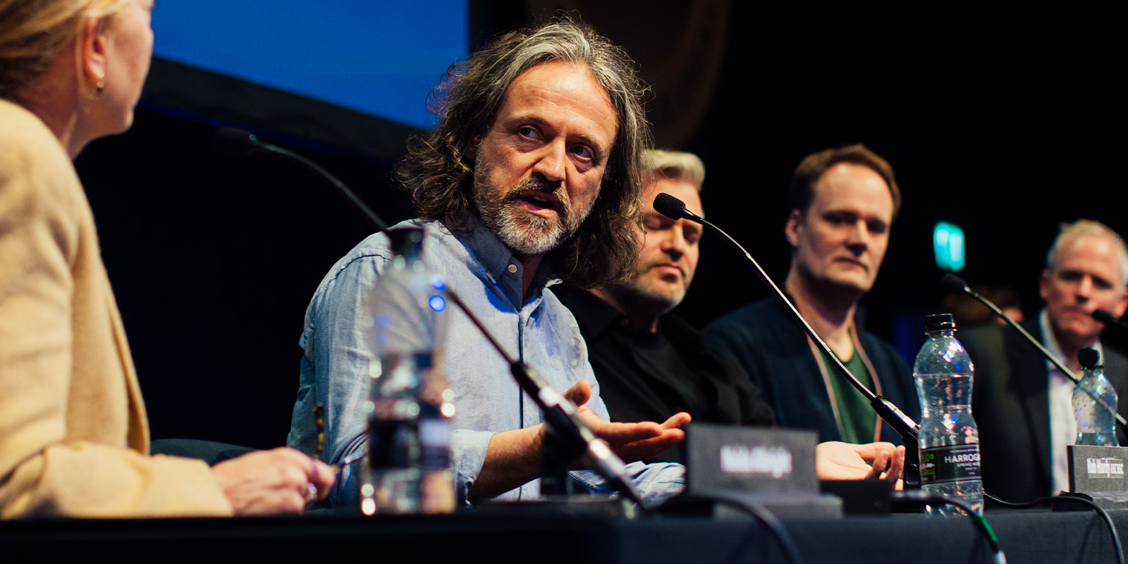 At a BSC Expo 2026 panel discussion, a man with long hair speaks whilst gesturing with his hands. Four others sit beside him, listening intently, with nameplates and water bottles arranged on the table in front of them.