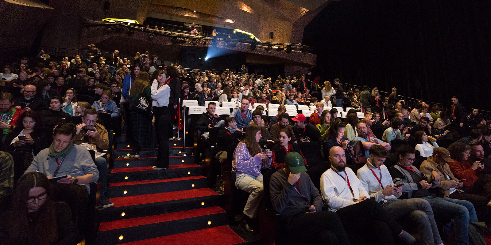 A large audience fills most seats in a dimly lit theatre at Camerimage 2026. Some attendees chat whilst others check their phones. The stage is out of view, with rows of red-carpeted stairs leading through the centre.