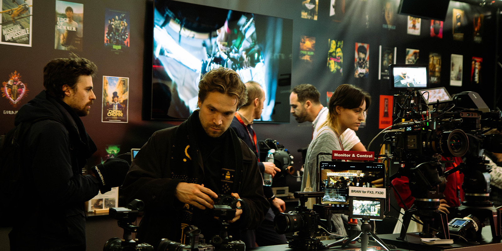 Several people examine professional cameras at an electronics or camera exhibition, possibly during Camerimage 2026. Posters and monitors displaying film scenes and camera footage are visible on the walls in the background.