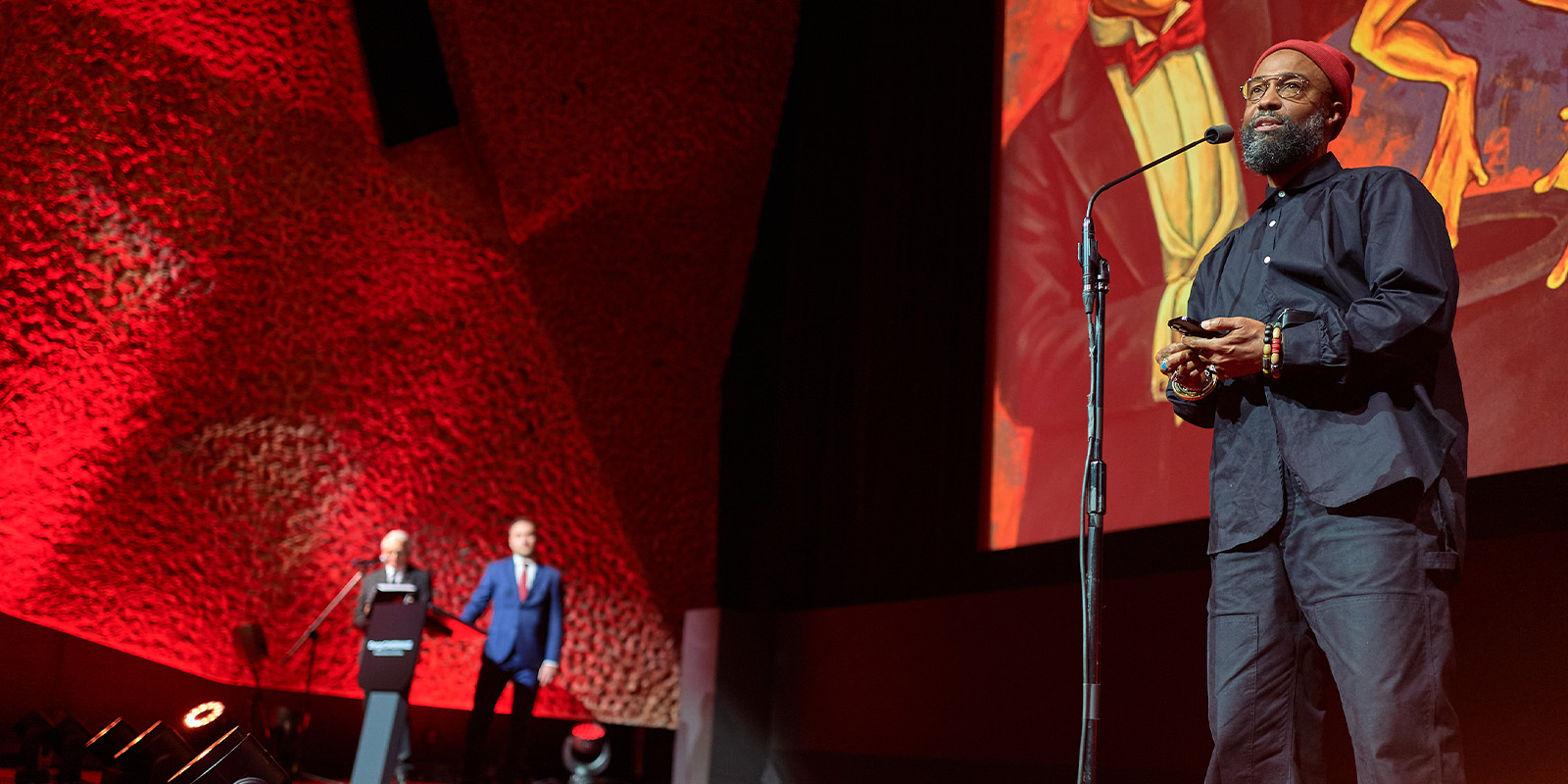 At Camerimage 2026, a man in dark clothing and a red hat speaks into a microphone on stage, holding an award. Two people stand at a lectern against a textured red wall, with a large portrait behind the speaker during the event review.