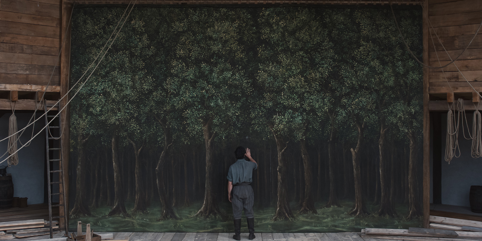 A person stands facing a large painted backdrop of a dense forest on a wooden stage, echoing the atmospheric visuals of Łukasz Żal, surrounded by ropes and wooden walls, with a ladder leaning against the left side.