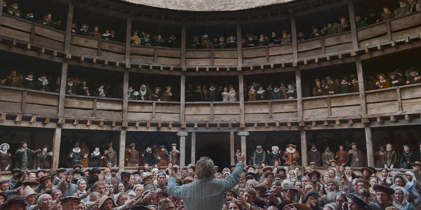 A person stands on a stage facing a large, attentive audience in an open-air, circular wooden theatre with three levels of seating, resembling an Elizabethan playhouse—evoking scenes from Hamnet, captured in the style of Łukasz Żal.