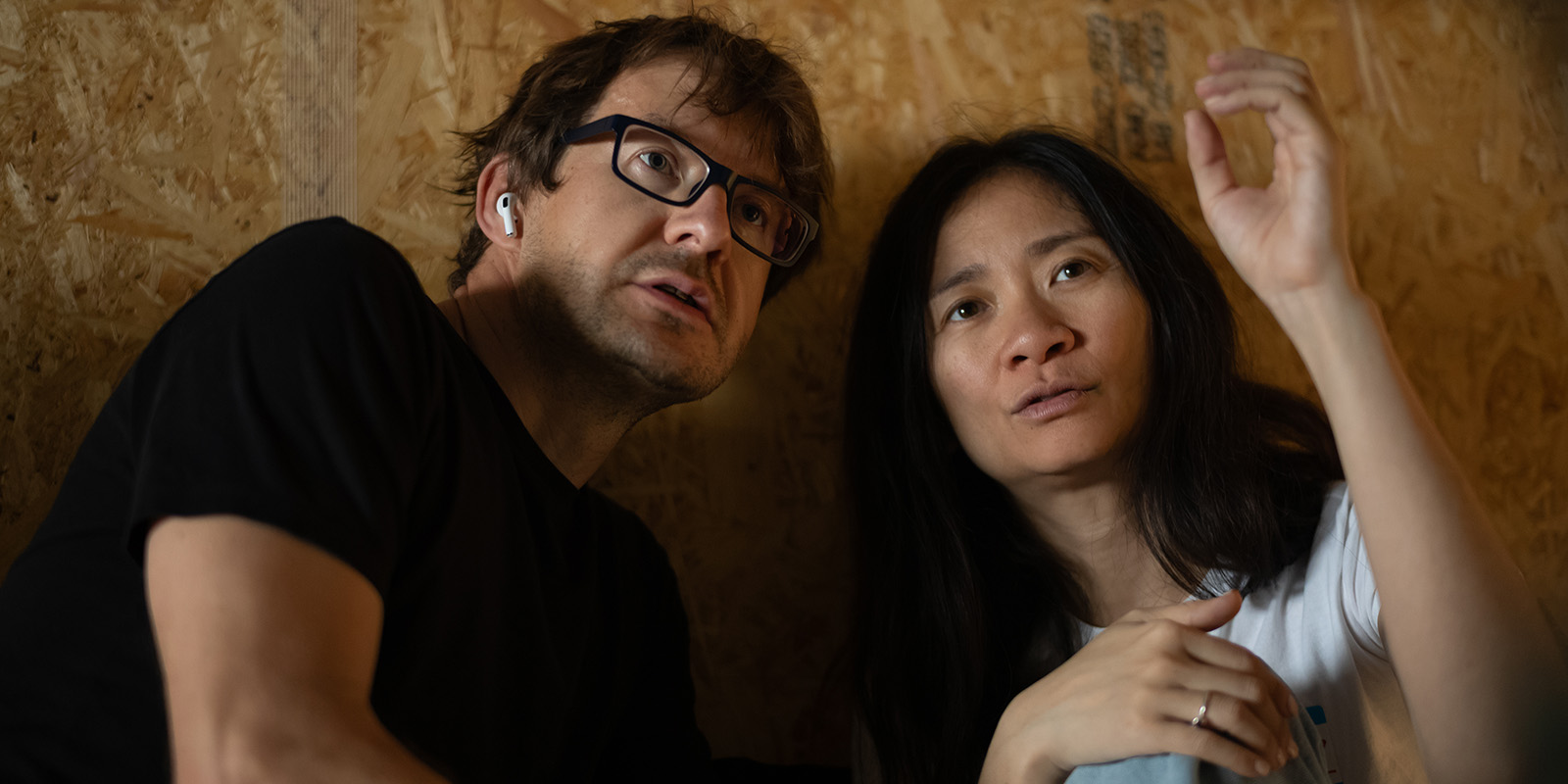 A man wearing glasses and earphones and a woman with long dark hair are sitting closely together indoors, looking slightly upwards with focused expressions, reminiscent of the cinematic style of Łukasz Żal, PSC. The background is a textured wood-panelled wall.