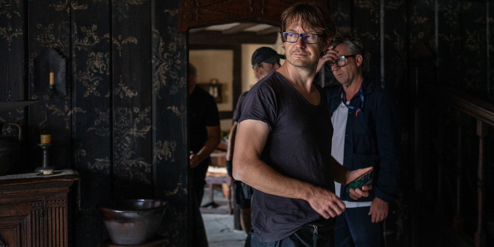 A man in glasses and a dark t-shirt stands indoors holding an object, with two other men and rustic wooden furniture visible in the background. The dimly lit setting, reminiscent of Łukasz Żal’s Hamnet style, features patterned wallpaper throughout.