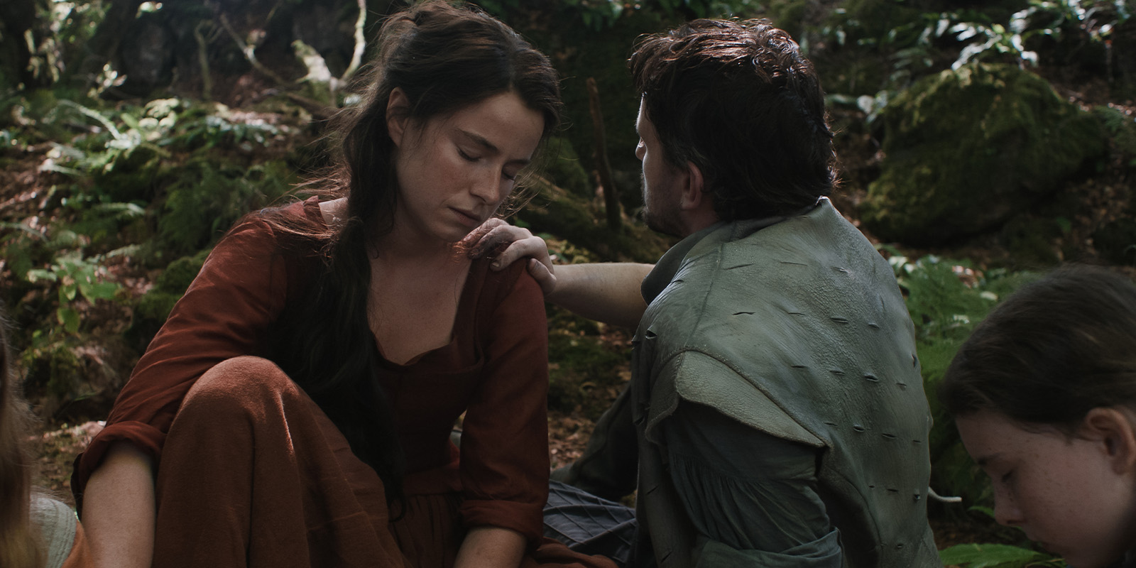 A woman in a red dress sits on the forest floor, eyes closed, while a man in a green tunic touches her shoulder—a scene from Hamnet, beautifully captured by Łukasz Żal, PSC. Dappled sunlight filters through lush greenery; another figure is partially visible.