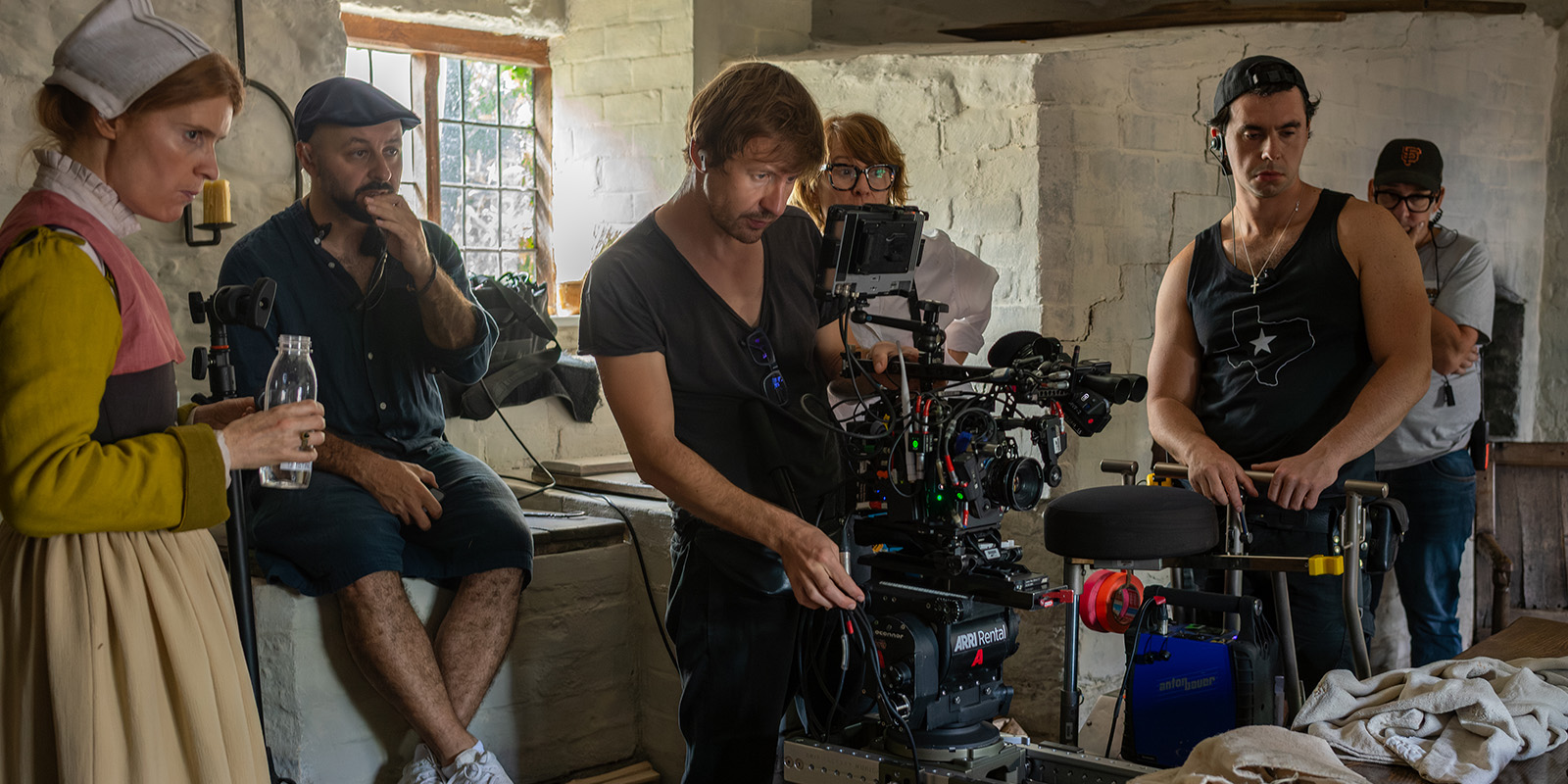 A film crew sets up a camera in a rustic room reminiscent of Hamnet. One person is dressed in period costume, whilst others, in casual clothing, focus on filmmaking equipment. Light, evocative of Łukasz Żal, PSC's style, streams from a window in the background.