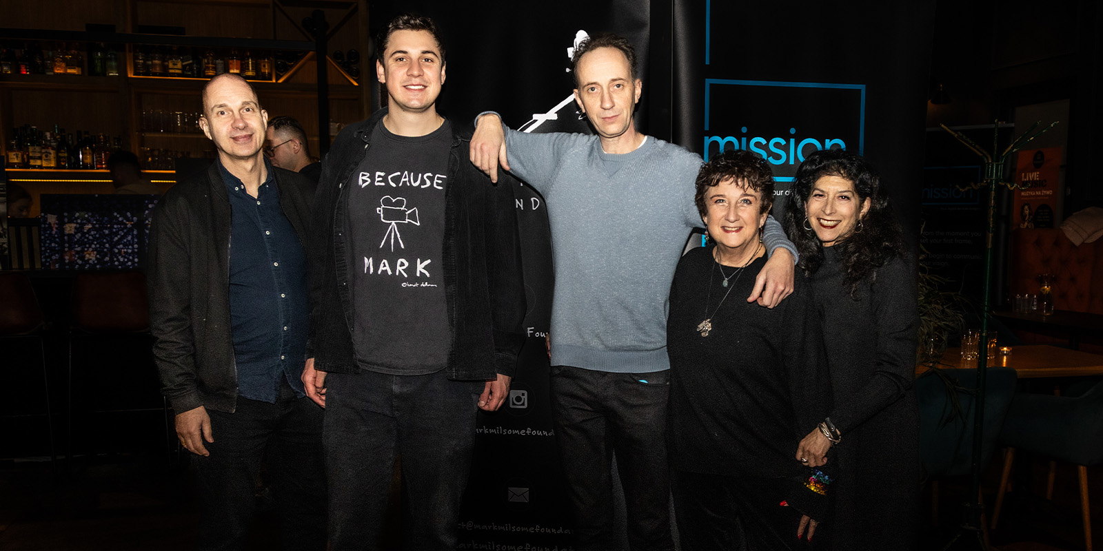 Five people stand together indoors in front of a dark backdrop at Camerimage 2026. Three men and two women pose for the camera, some with arms round each other, with a bar and event review banners visible in the background.