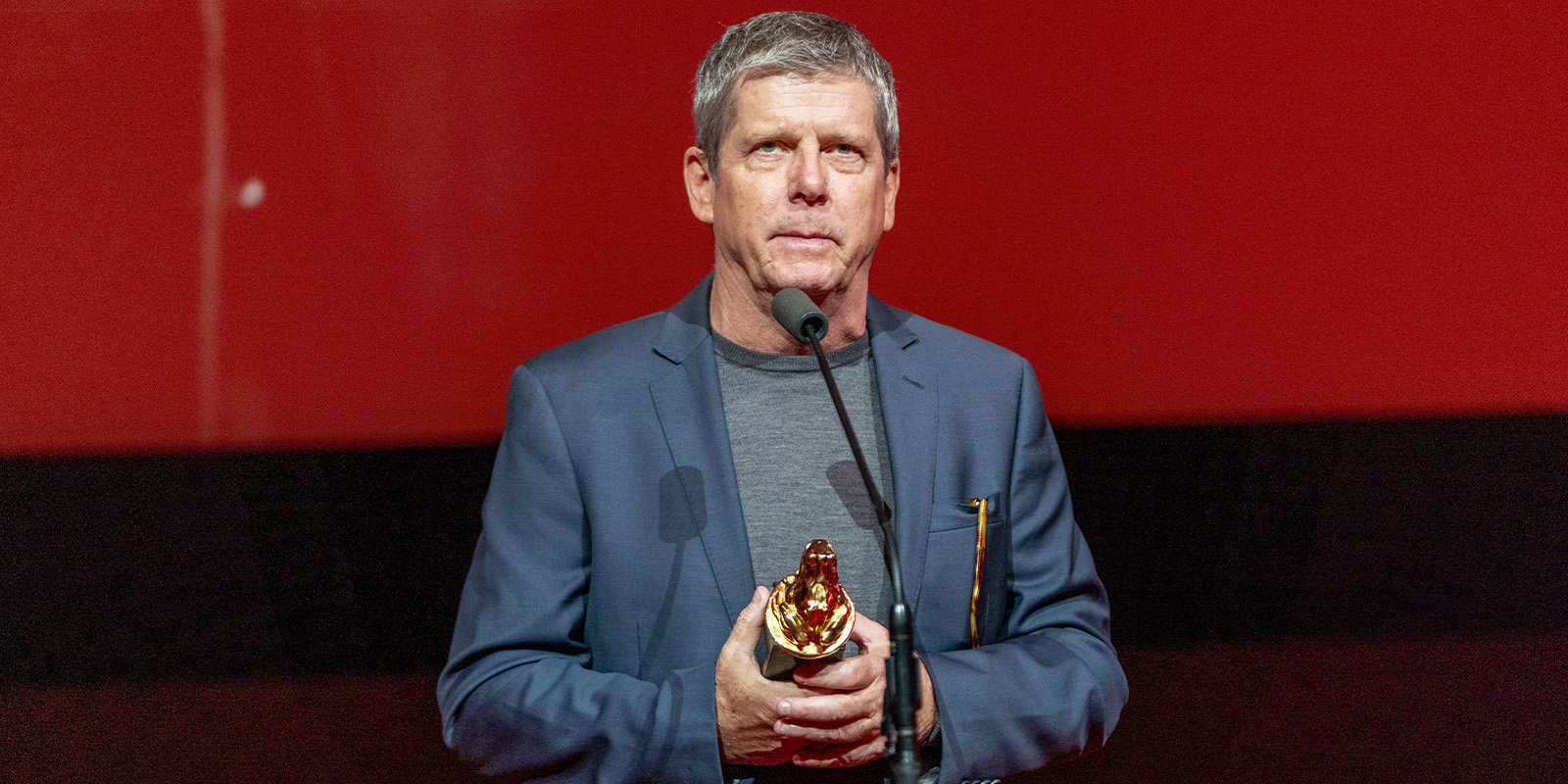 A man in a blue blazer and grey shirt stands at a microphone holding a golden bear-shaped trophy against a red background, celebrating his achievement at Camerimage 2026. Event review highlights his inspiring moment on stage.