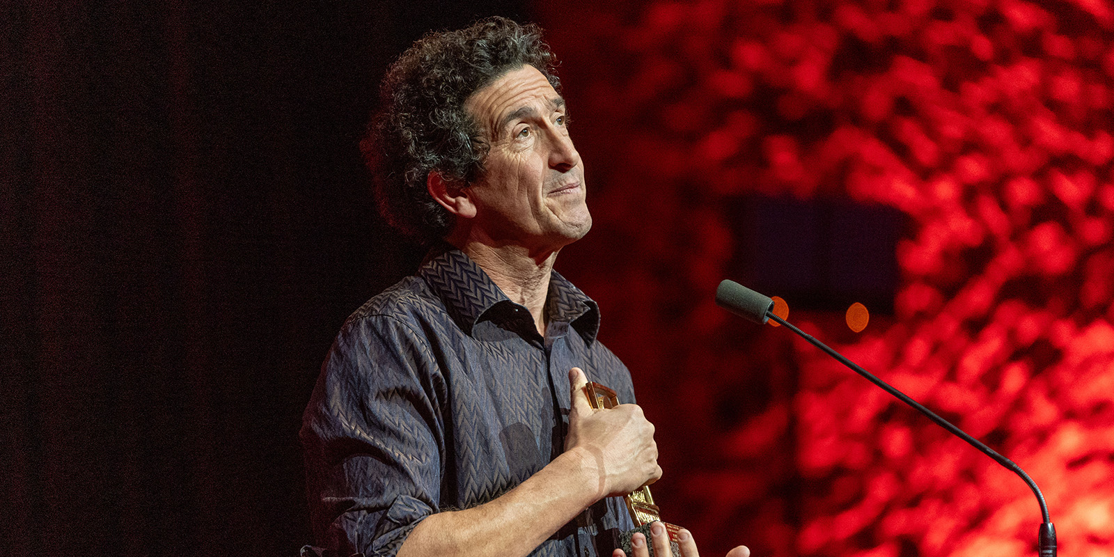 A man stands at a microphone on stage at Camerimage 2026, holding an award to his chest. Against a red, textured background, he appears to be speaking or reflecting—capturing a key moment perfect for any event review.