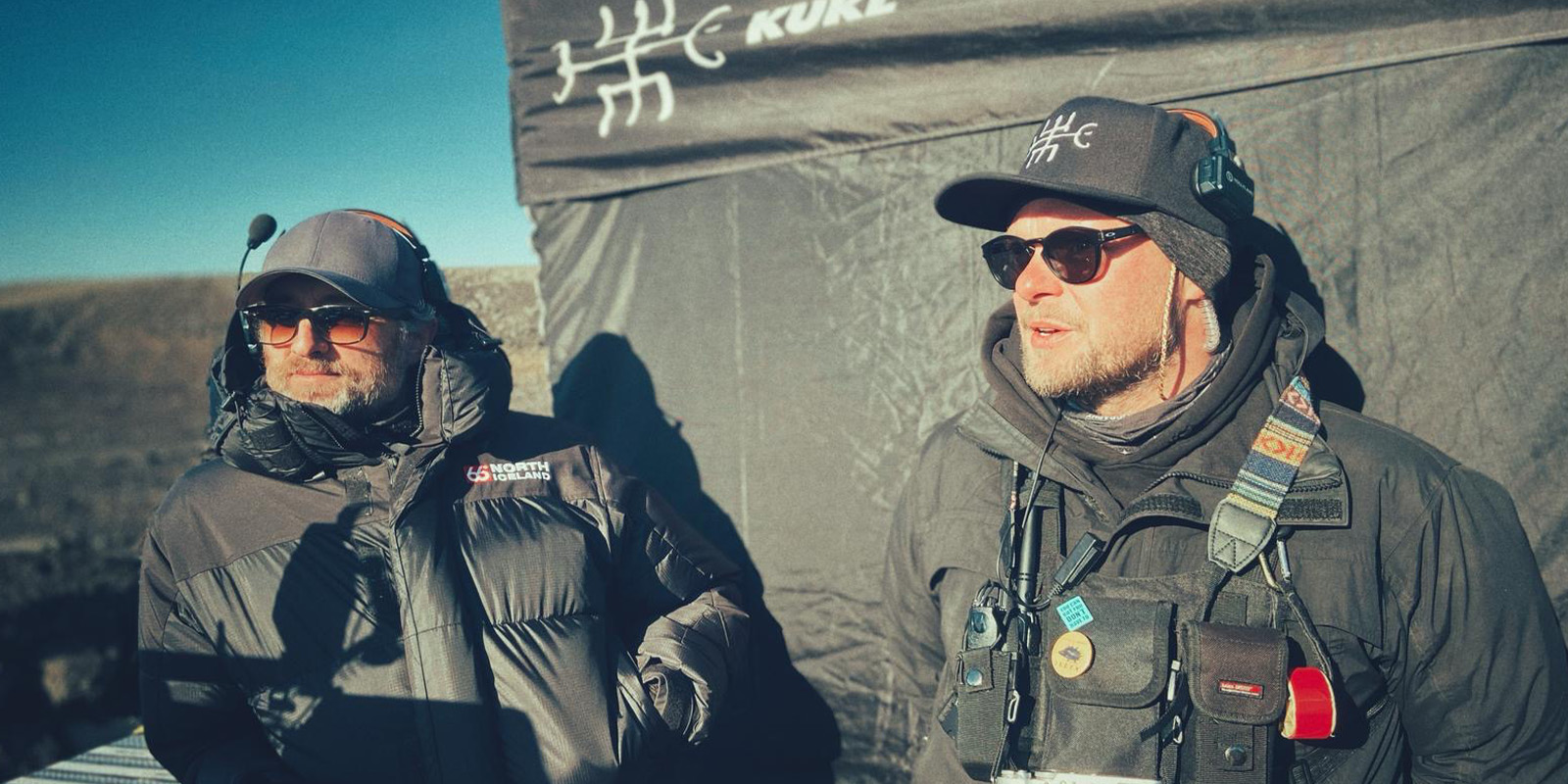 Two men wearing sunglasses, hats, and headsets stand outdoors near a tent with gear attached to their jackets. The tent has a logo and the word KURZ printed on it. Ollie Downey appears to be part of the scene on this sunny day under a clear blue sky.