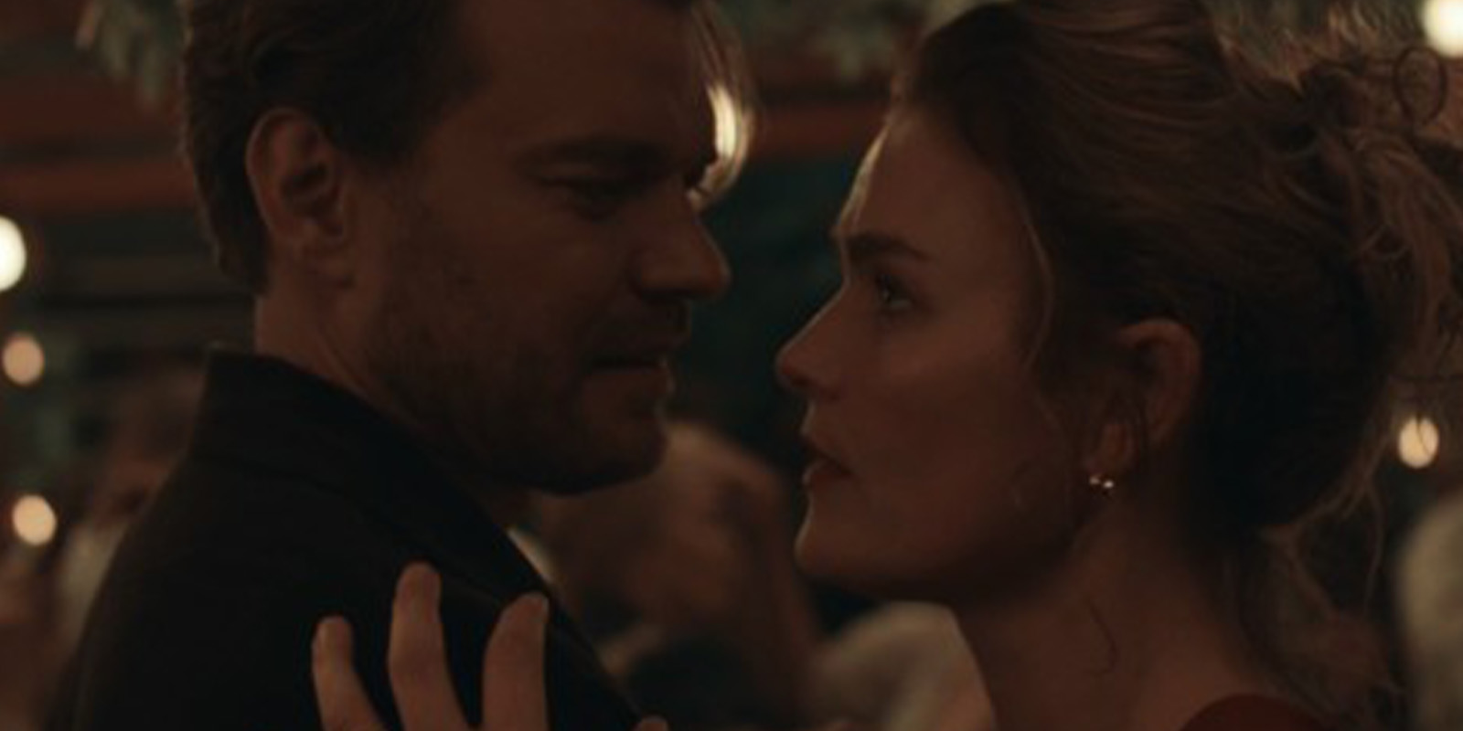 A man and a woman stand close together at a dimly lit event, gazing into each other's eyes. The background is blurred with lights and other guests, capturing the intimate atmosphere featured in Ollie Downey's work on The Fires.