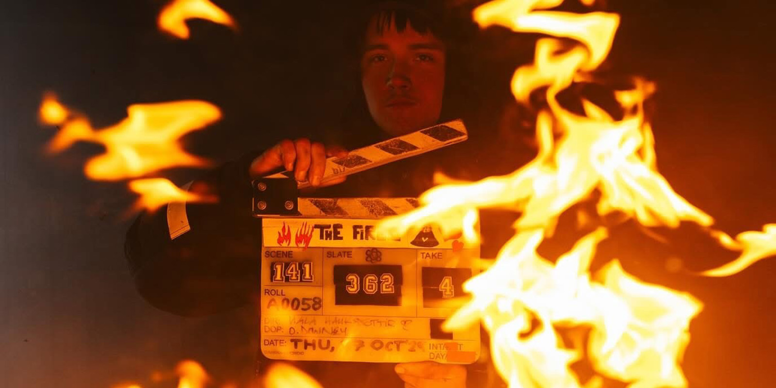 A person holds a film clapperboard surrounded by large orange flames. The clapperboard, marked with "The Fires" and Ollie Downey, is partially obscured by the fire, whilst the person's face is dimly visible in the background.