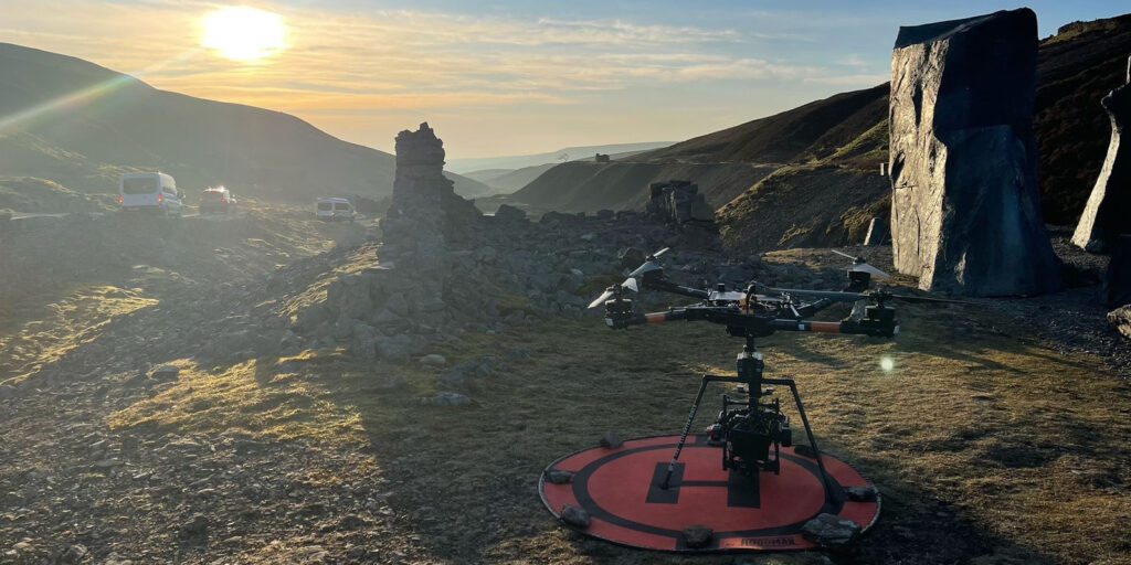 A large drone sits on a red landing pad in a rocky outdoor area at sunset, with hills in the background, ruined stone structures, parked vehicles, and a winding road on the left.