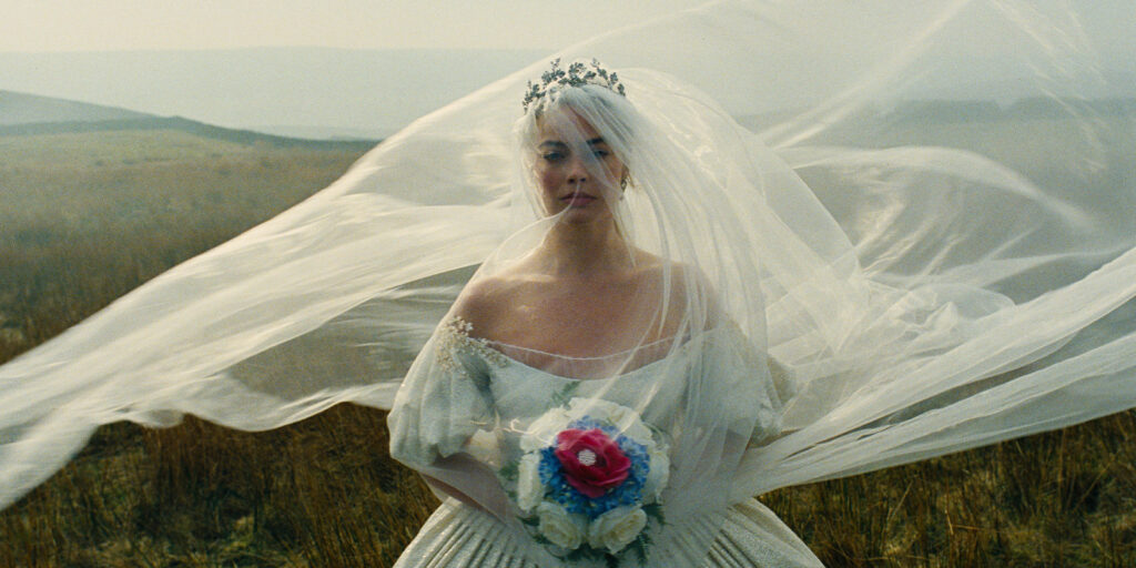 A person in a white wedding dress and veil stands outdoors, holding a bouquet of colourful flowers. The veil is billowing in the wind, and a landscape of fields and hills is visible in the background.