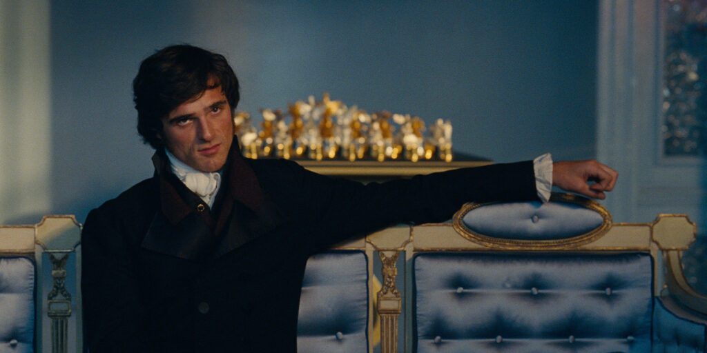 A man in 19th-century style clothing sits on a blue tufted sofa with gold trim, resting one arm on the backrest. There are small golden figurines on a shelf in the background.
