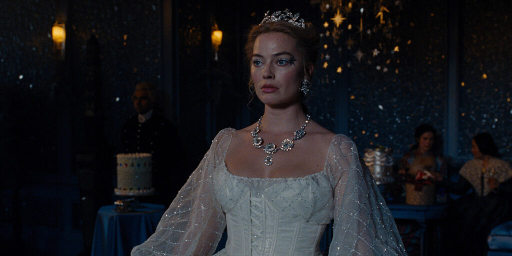 A woman in a white, beaded gown and tiara stands in a dimly lit room decorated with star-shaped lights. She wears a large jewelled necklace. In the background, people sit at tables with cakes and candles.