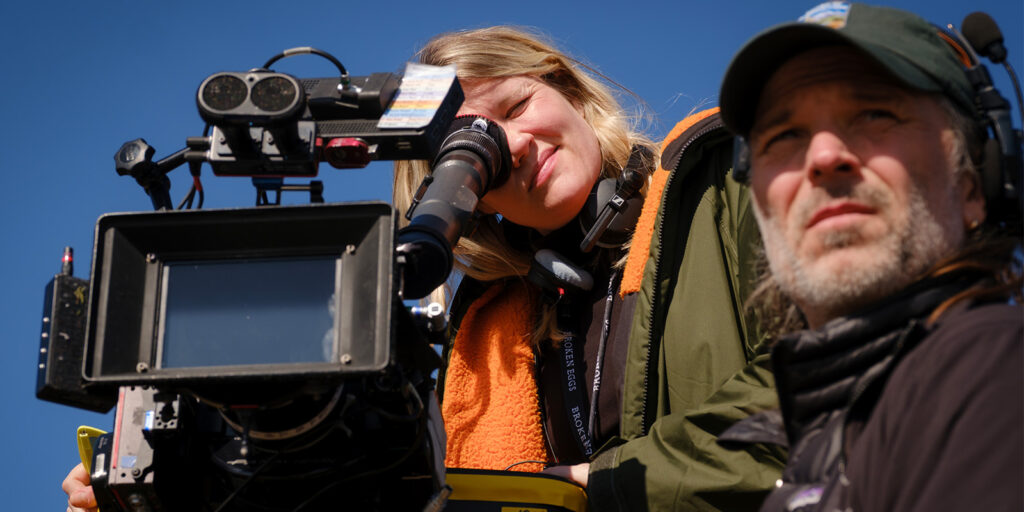 A woman looks through the viewfinder of a professional film camera while a man wearing a headset stands beside her, both outdoors under a clear blue sky.