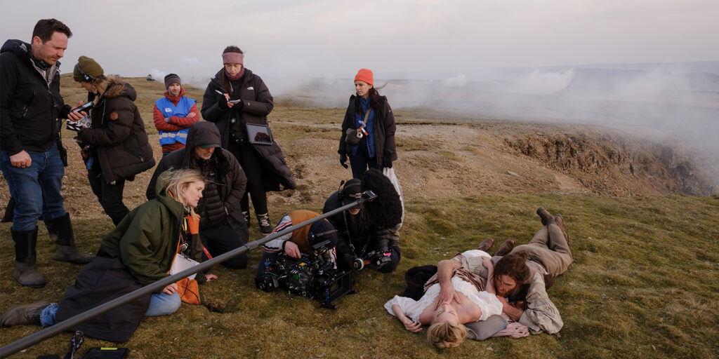 A film crew is working outdoors on a grassy plain with smoke or steam in the background. Two actors lie on the ground performing a scene whilst crew members operate cameras and equipment around them.