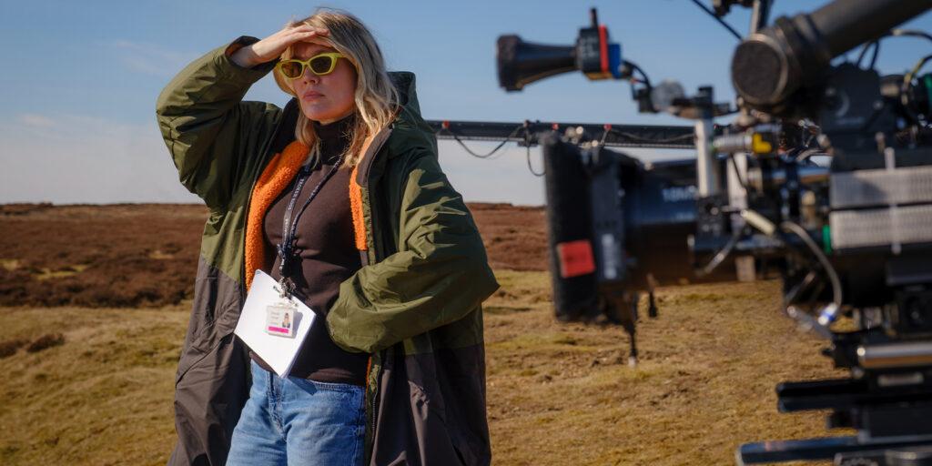 A person wearing sunglasses and a long coat stands outdoors on a grassy field, shielding their eyes from the sun. A camera and filming equipment are visible in the foreground. The person has a lanyard ID badge.