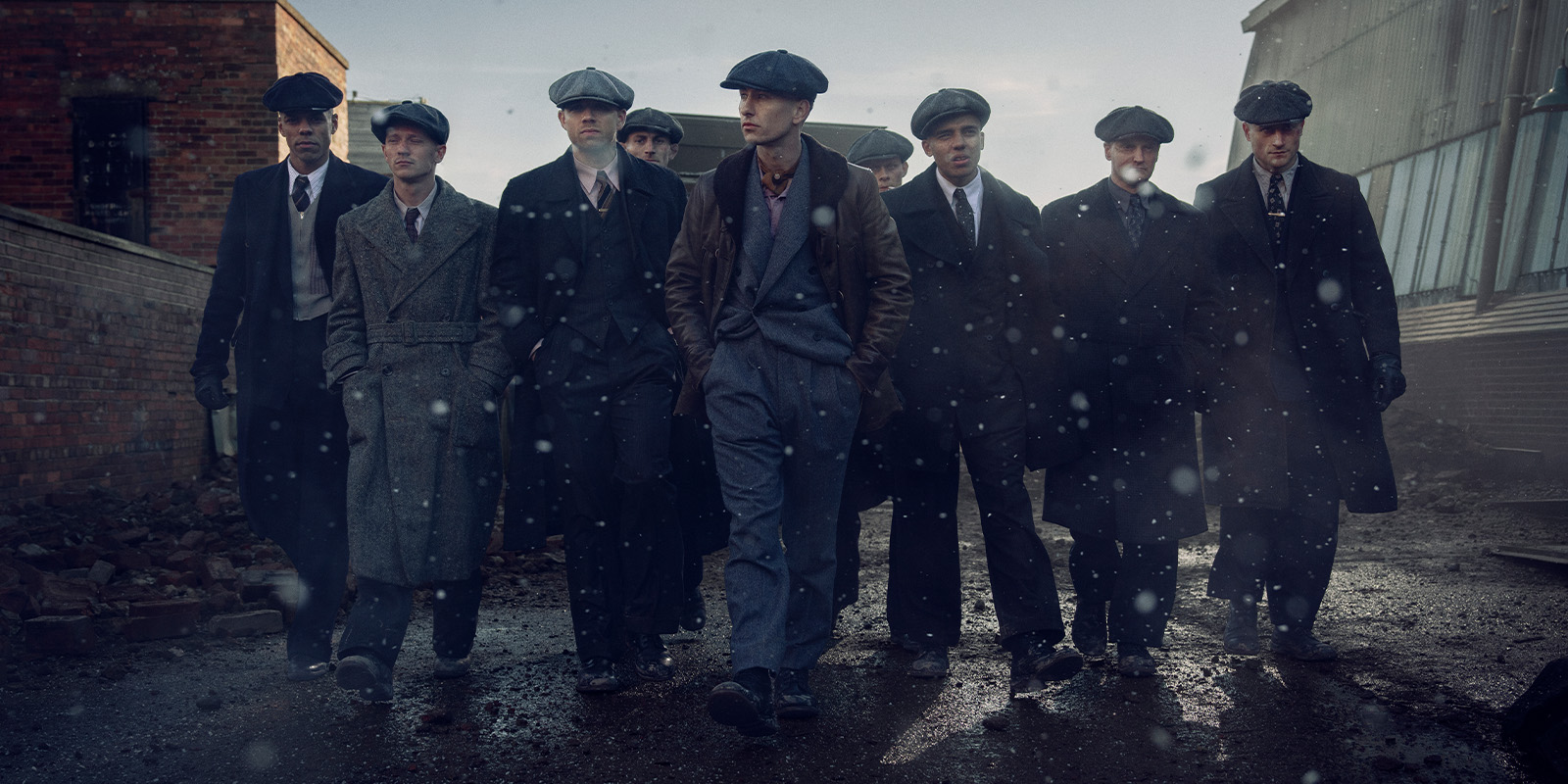 A group of nine men in early 20th-century attire, including flat caps and long coats, walk together on a wet, debris-strewn street in an industrial setting, with light snow or rain falling.