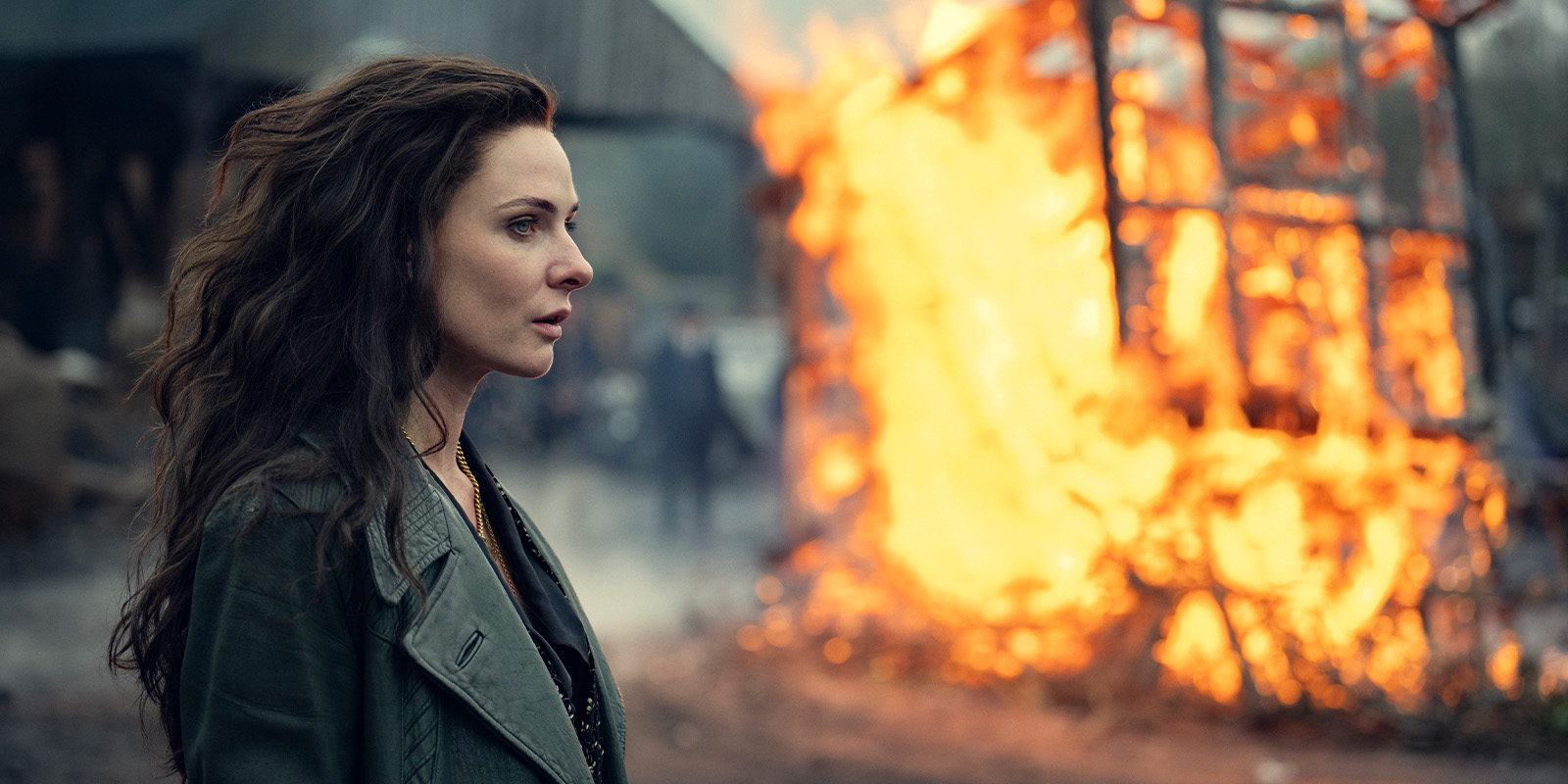 A woman with long, wavy brown hair and a green jacket stands in profile, looking serious, whilst a large wooden structure burns intensely in the background, creating smoke and bright flames.