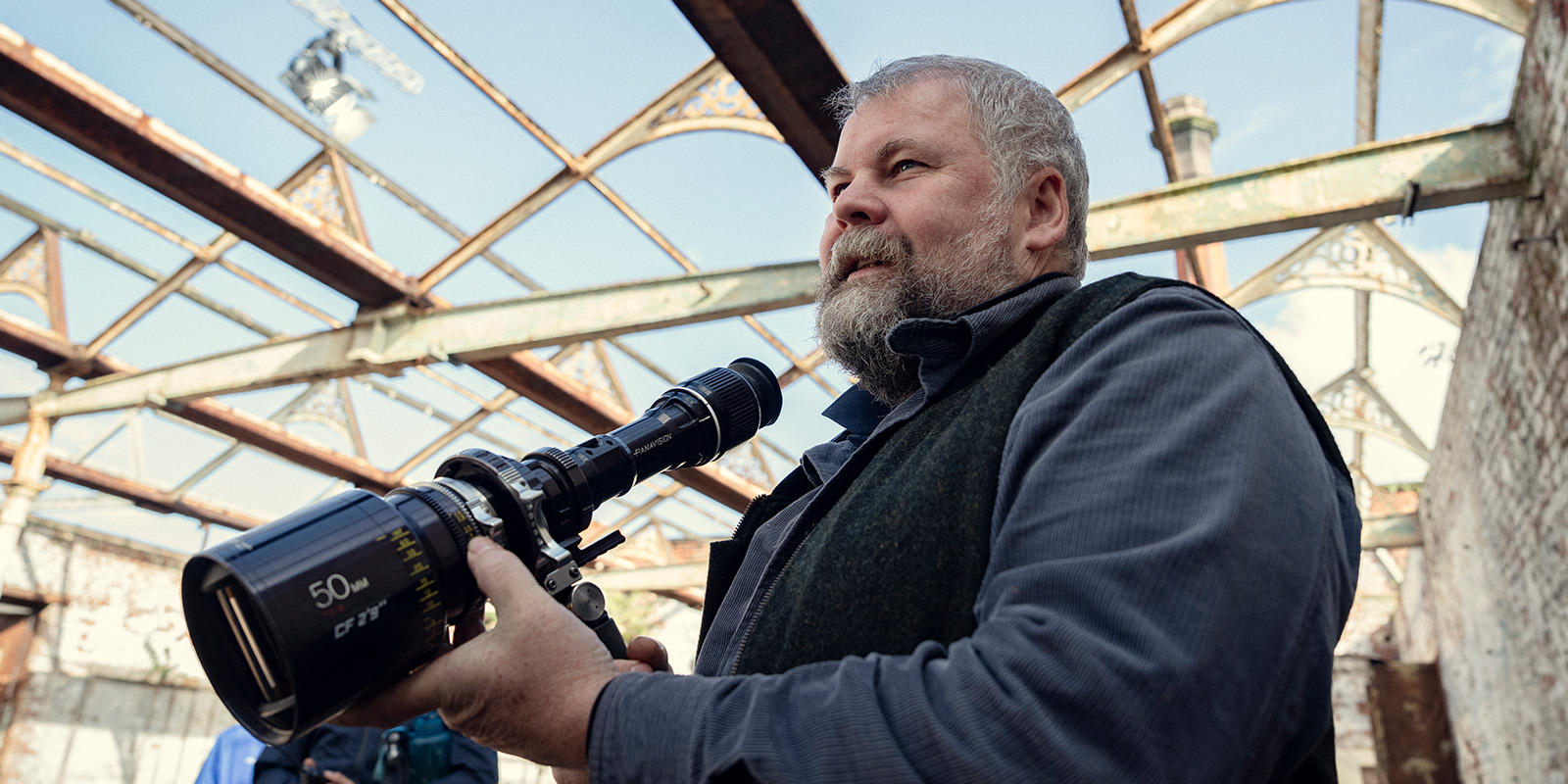 A bearded man in a blue shirt holds a large camera lens inside a partially constructed or dilapidated building with exposed beams and no roof.