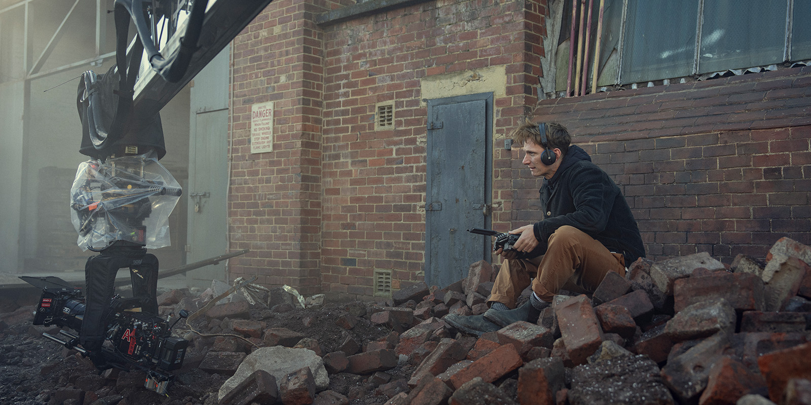 A person wearing headphones sits on a pile of bricks next to a brick building, holding equipment. A film camera mounted on a crane is positioned nearby, with another person operating it under a plastic cover.