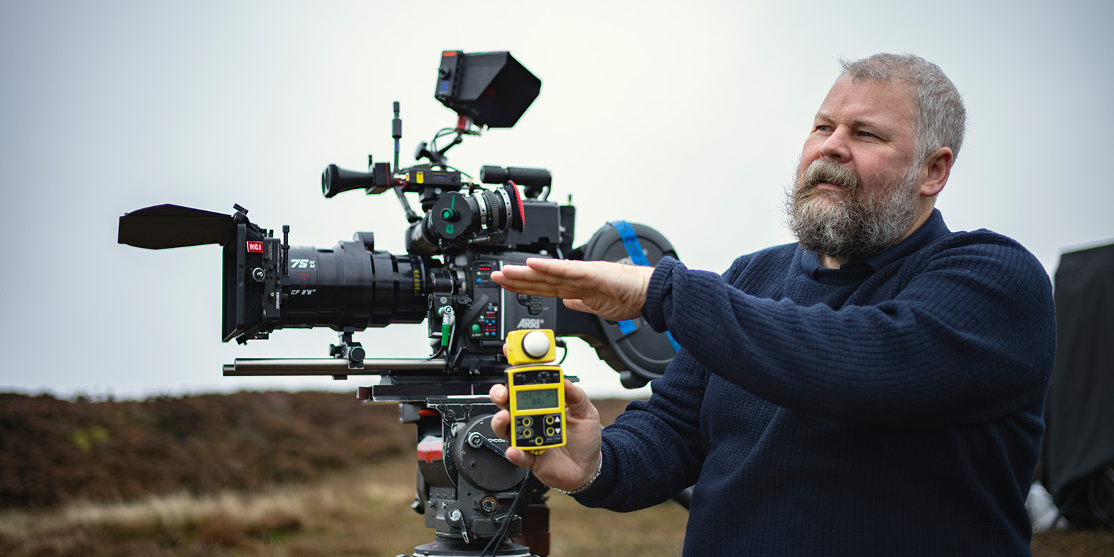 A bearded man outdoors stands next to a professional video camera on a tripod, holding a yellow and black device in one hand and gesturing with the other. The background is slightly blurred with cloudy sky.