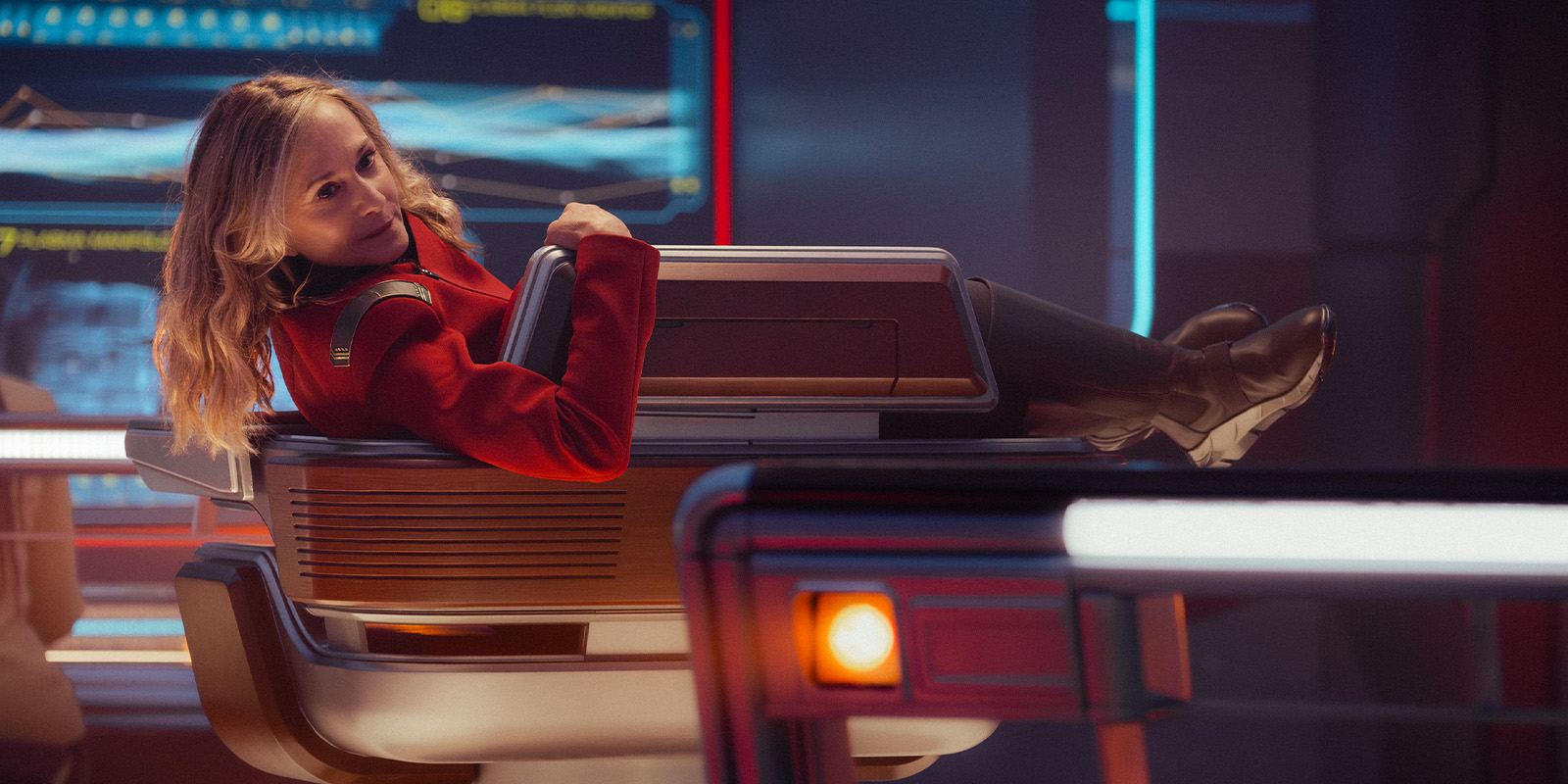 A person in a red uniform reclines sideways on a futuristic chair in a brightly lit control room with large digital screens and illuminated panels in the background.