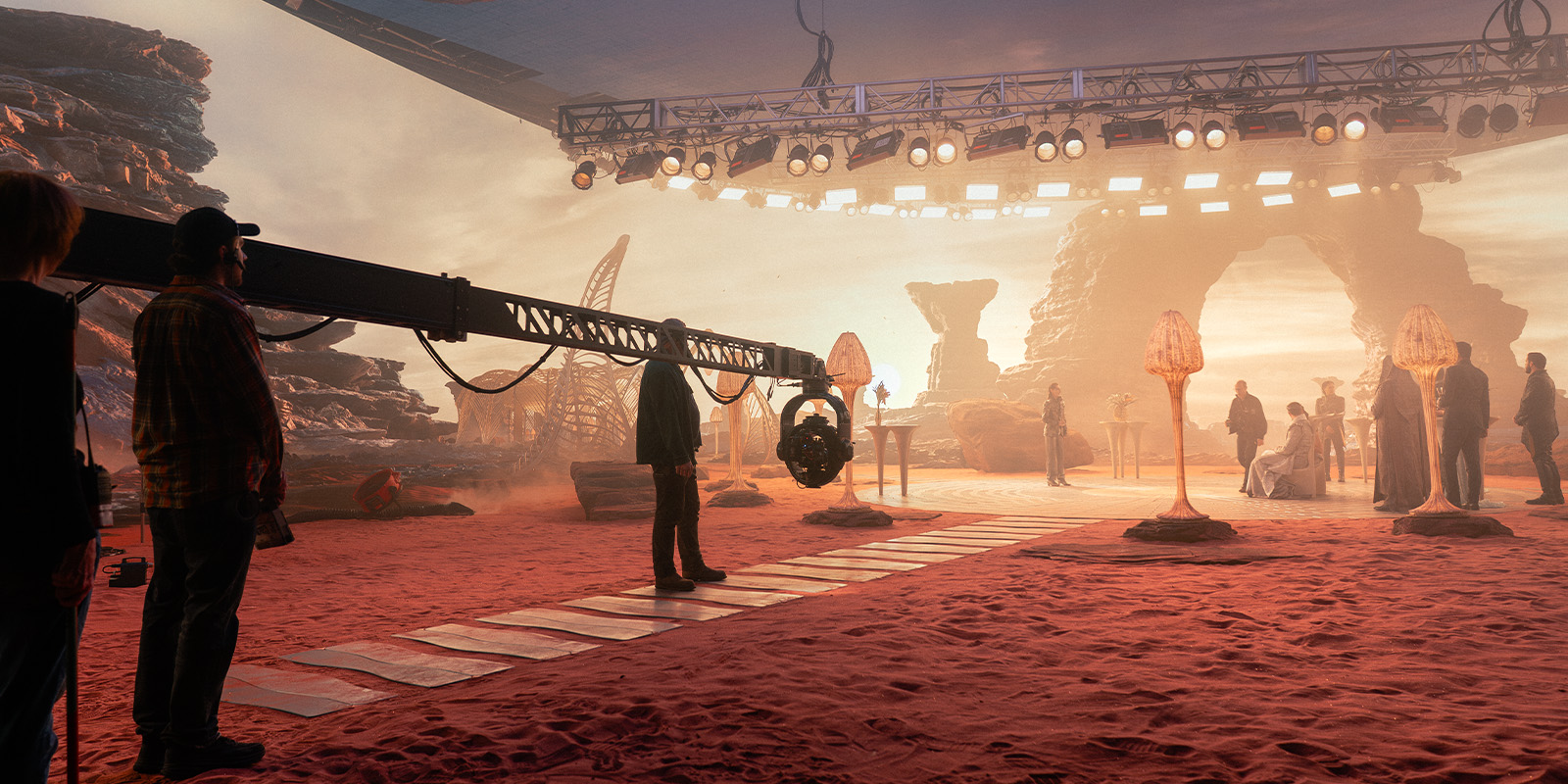 A film crew is shooting a scene on a set designed to look like a rocky desert with artificial rocks and orange lighting. Crew members stand near a camera crane, and actors in costume are filming in the background.