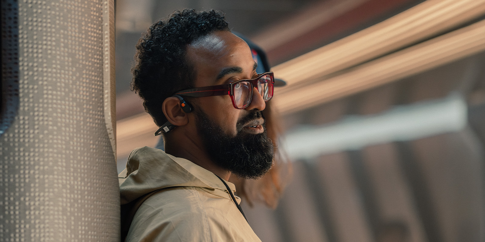 A man with a beard and glasses, wearing a tan jacket and earphones, leans against a patterned wall in an indoor setting with blurred background lighting.