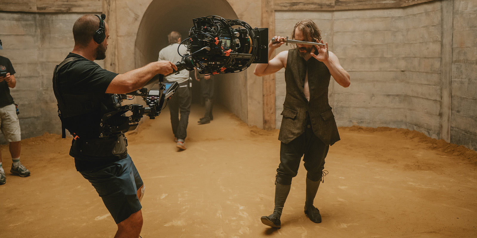 A camera operator films an actor holding a sword in front of his face on a sandy set with concrete walls. Other crew members are visible in the background.
