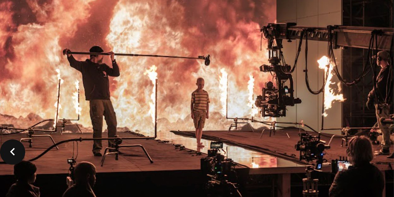 Film crew records a young person standing on a set with fire and a backdrop of flames. Multiple cameras, lights, and boom microphones are visible, capturing the scene from different angles.