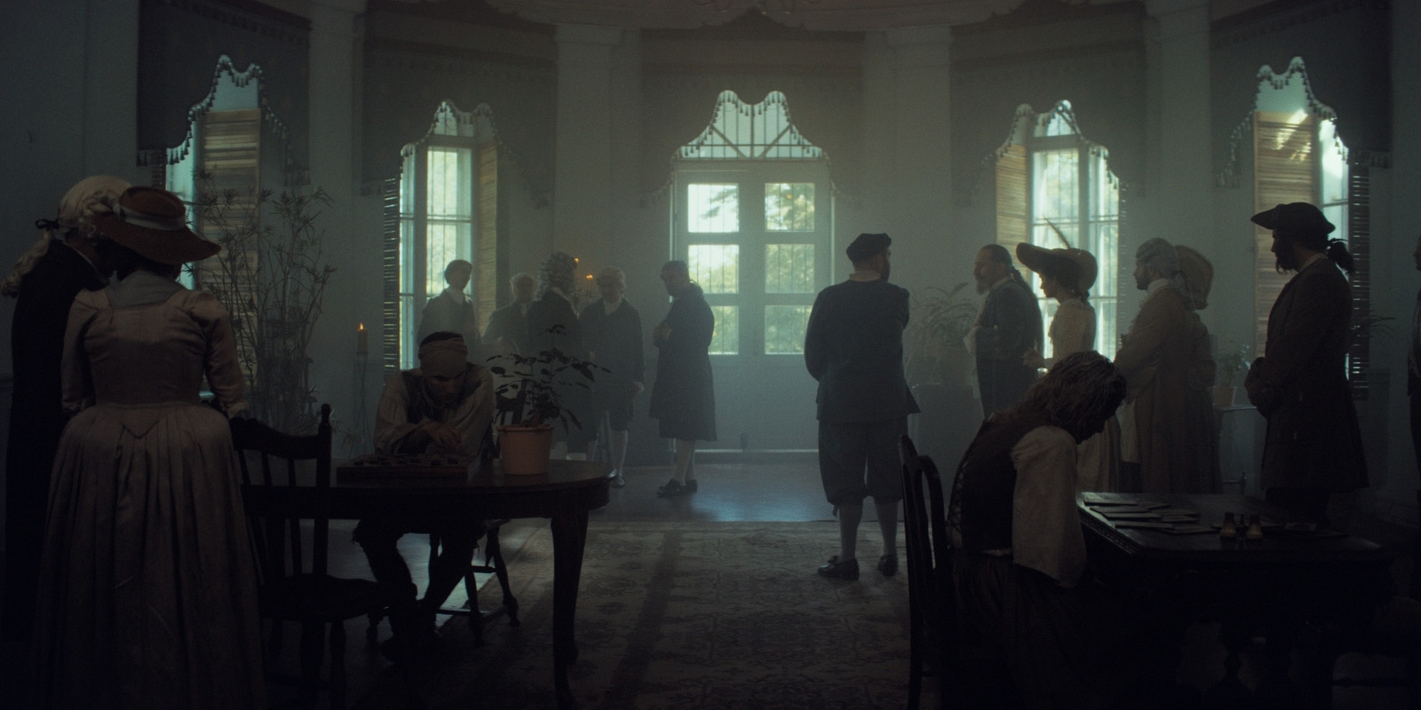 A group of people in period clothing stand and sit in a dimly lit, vintage room with large windows, ornate decor, and antique furniture. Some sit at tables whilst others converse or look out of the windows.