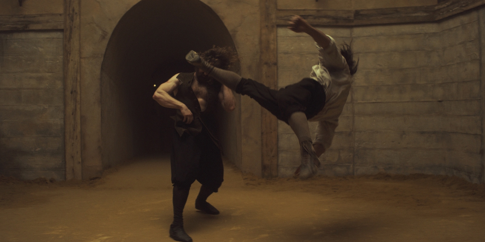 Two people are fighting in a dimly lit corridor, with one person mid-air delivering a flying kick towards the other person's head. The walls are rough stone, and the ground is covered in sand or dust.