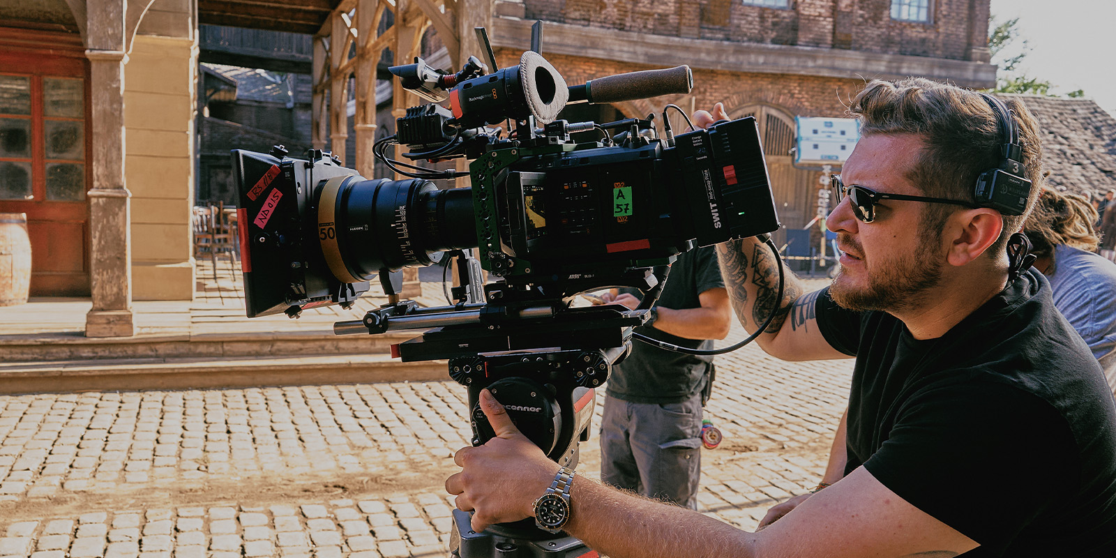 A man wearing sunglasses and headphones operates a professional video camera on a tripod outdoors in a setting with cobbled pavement and rustic buildings.
