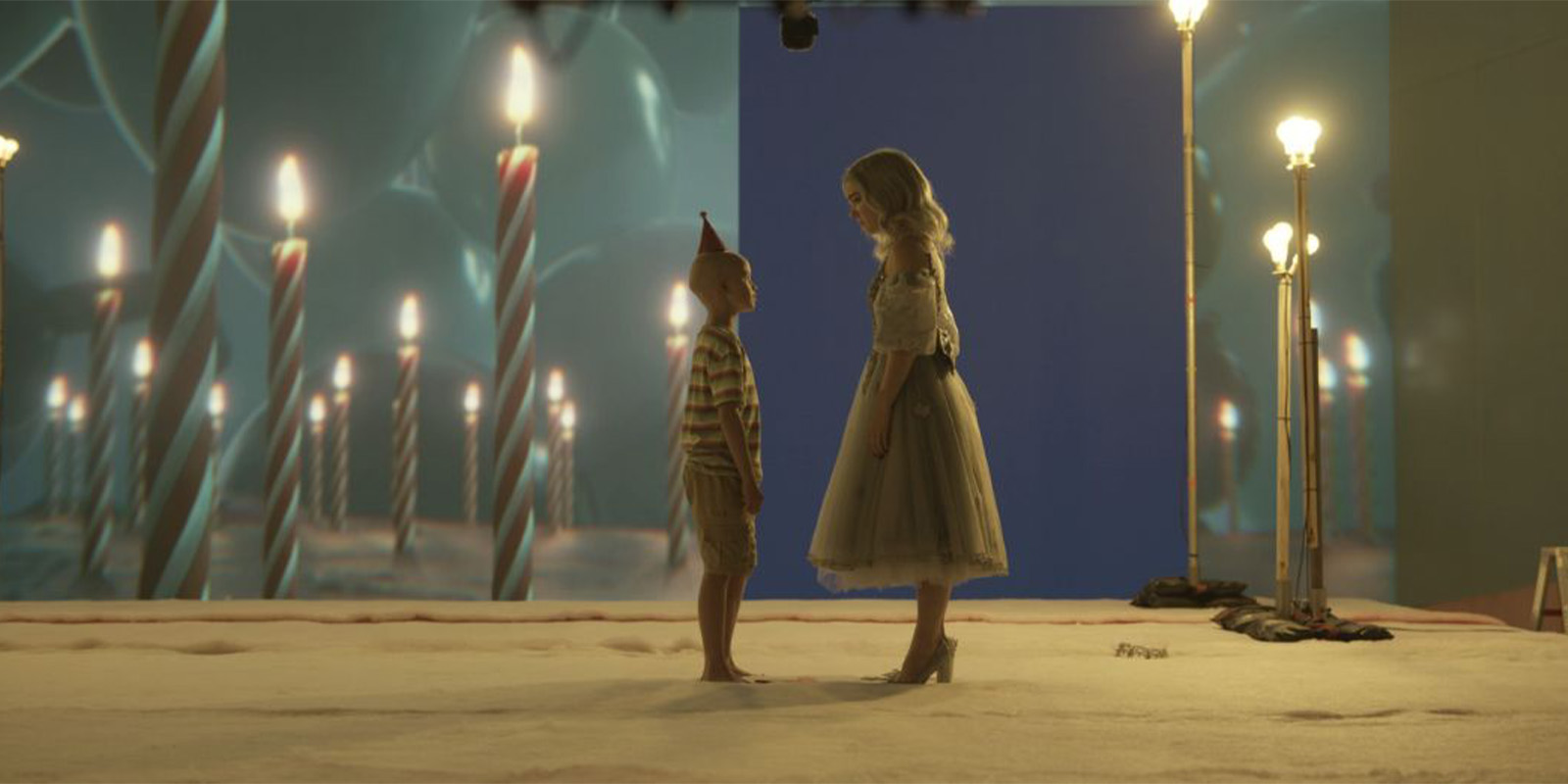 A young boy in a striped shirt and party hat stands facing a woman in a dress and heels; they are on a set with a backdrop featuring large birthday candles and surrounded by old-fashioned lamp posts.