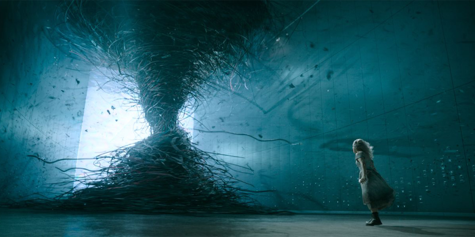 A person in a long coat stands in a vast, dimly lit room, facing a large swirling vortex made of dark, twisting wires or tendrils extending from floor to ceiling.