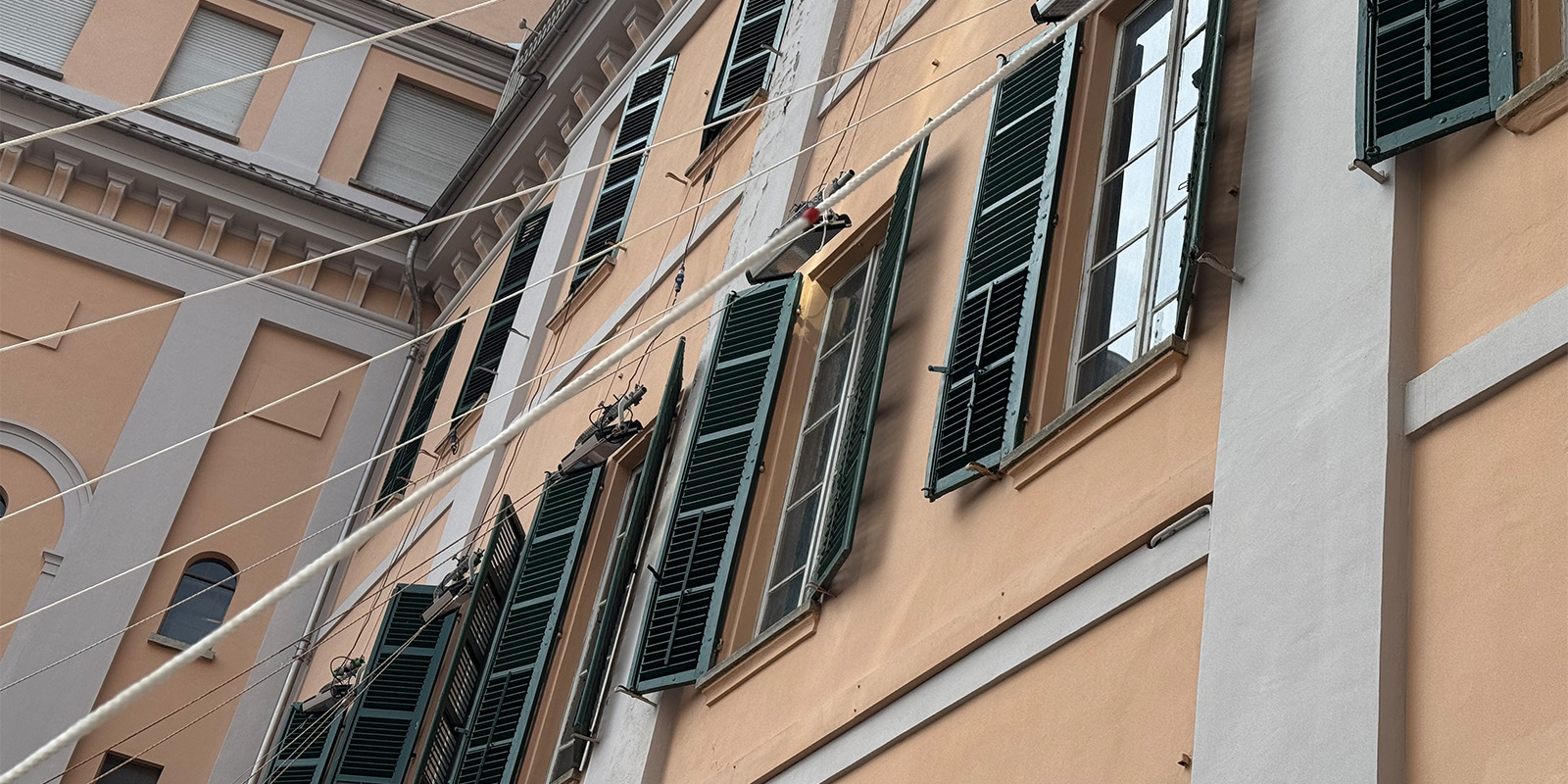 A peach-coloured building facade with green shutters, some open and some closed, on tall windows. Several white cables run diagonally across the scene in front of the building.