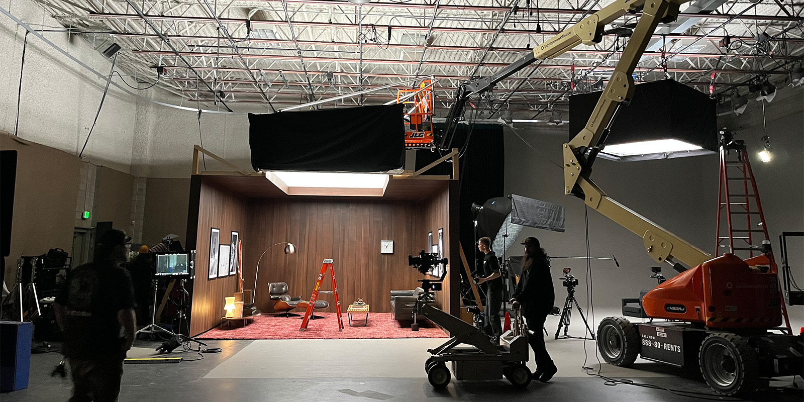 A film set inside a studio, featuring a furnished living room with wood-panelled walls, cameras, lights, tripods, large equipment, crew members, and a red ladder in the centre of the set.