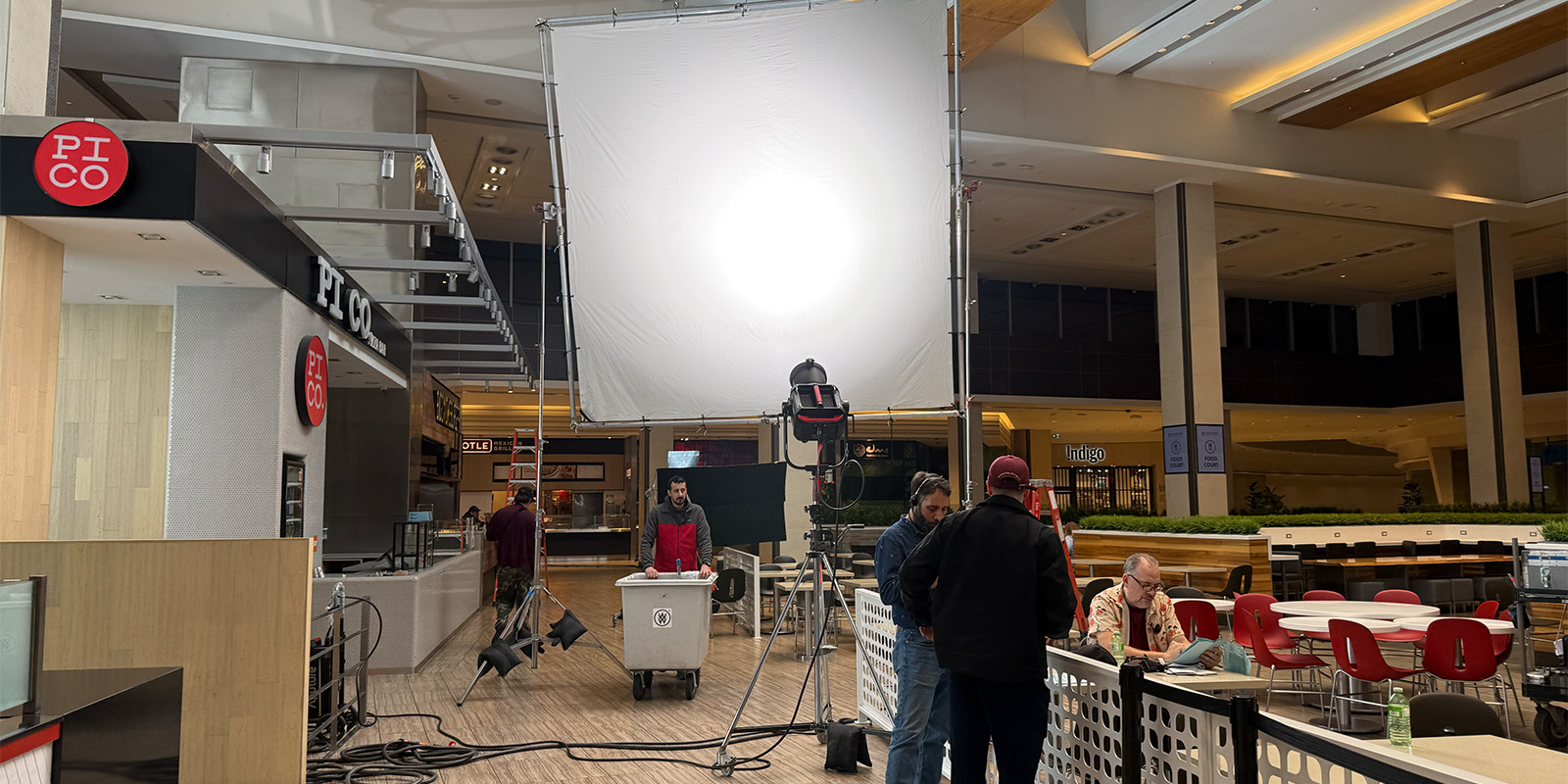 A film crew sets up lighting and equipment in a food court area, with several crew members working near a large white diffusion screen and camera gear. Some people are seated at tables in the foreground.