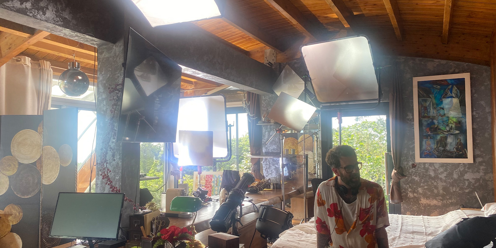 A cluttered room with large photography lights, a man with glasses and headphones, a wooden ceiling, multiple artworks, and windows letting in natural light. Various electronic devices and decorations are on tables.