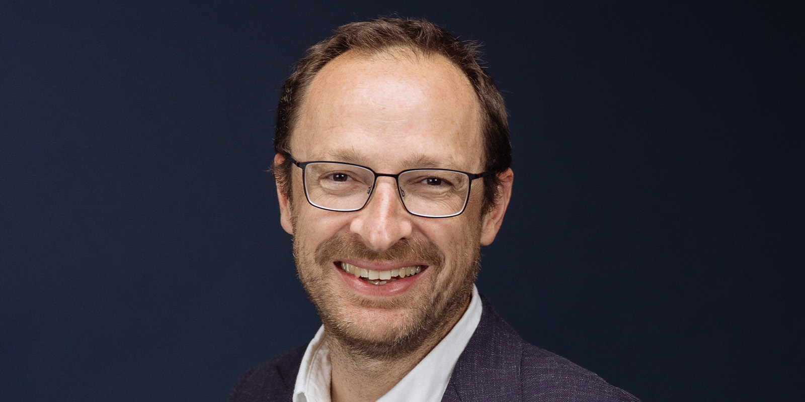 A man with short brown hair, glasses, and a beard is smiling at the camera. He is wearing a dark jacket over a white shirt and is posed against a plain dark background.
