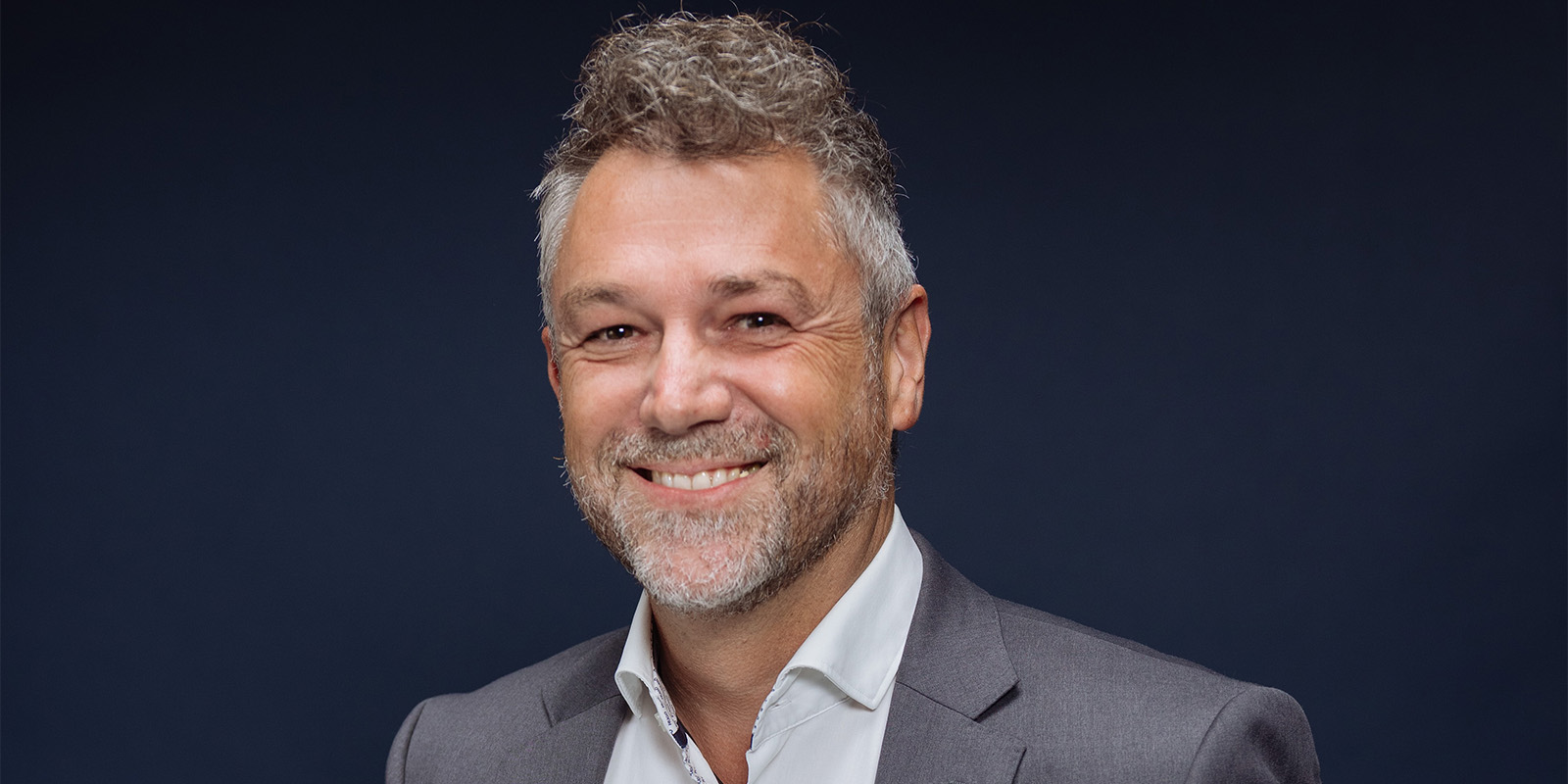 A man with short curly grey hair and a beard, wearing a grey suit jacket and white shirt, smiles at the camera against a plain dark background.