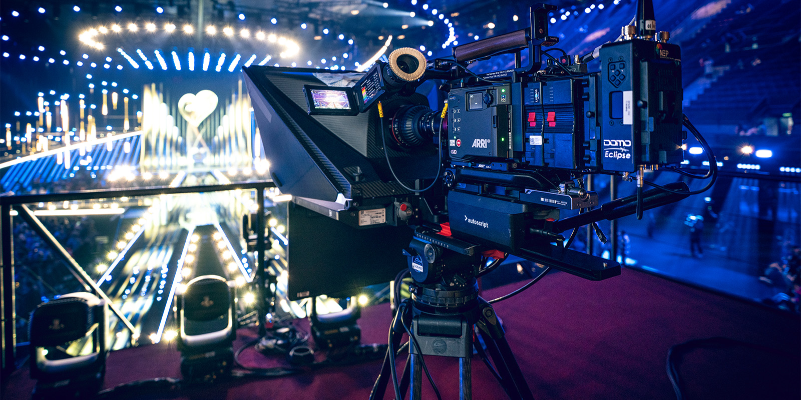 A professional video camera is set up at a live event with bright stage lights and an illuminated audience area visible in the background. The camera is focused on the stage.